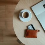 木のテーブルに置いたコーヒーとノートパソコン、カードを差した茶色の革小銭入れ